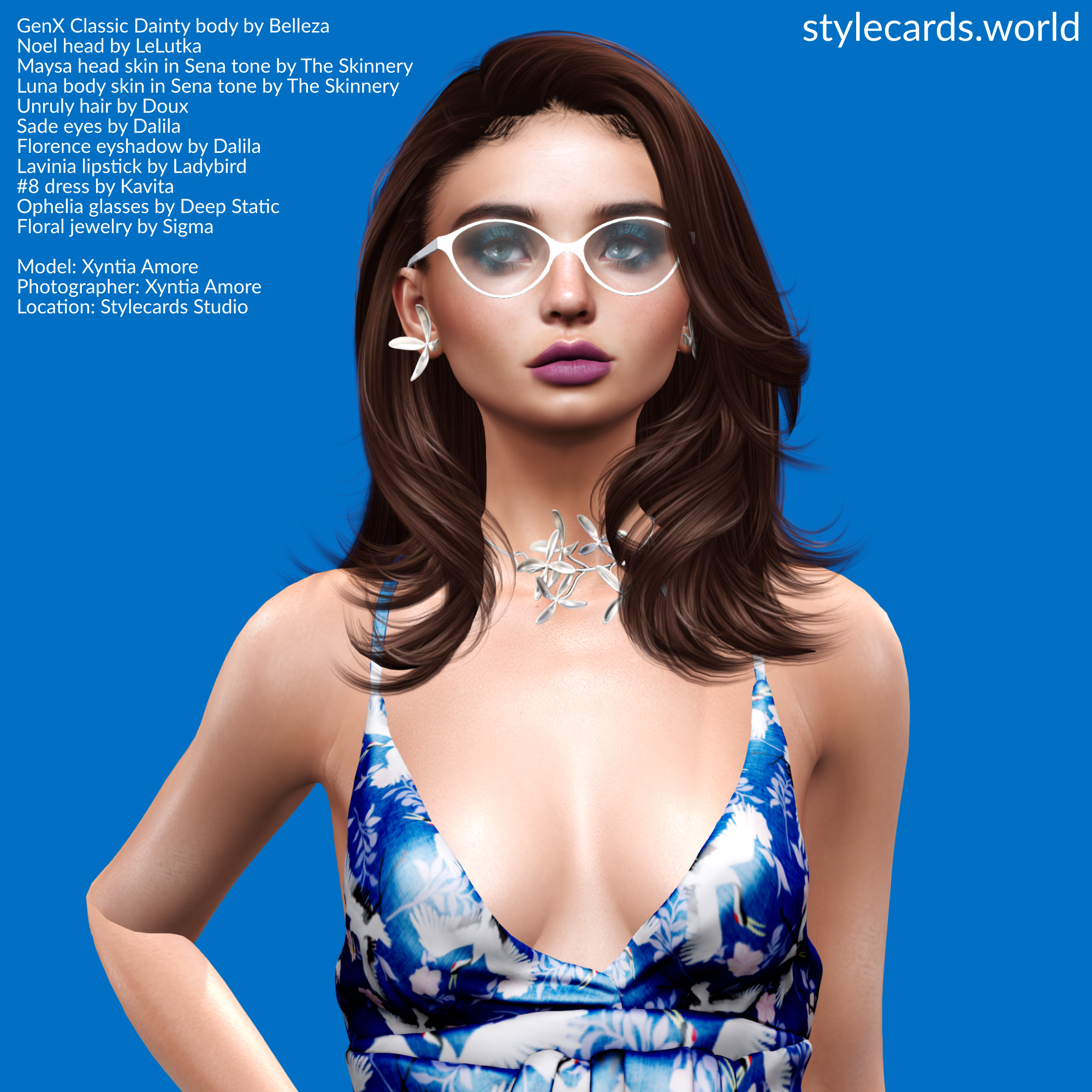 A headshot of Xyntia Amore has her casually posing in front of a blue backdrop while wearing a blue-and-white floral patterned dress. She has long brown hair, white framed eyeglasses, dark blue eyeshadow and purple-pink lipstick. Additionally, she is wearing a pair of silver floral-themed earrings and a matching necklace from Sigma.