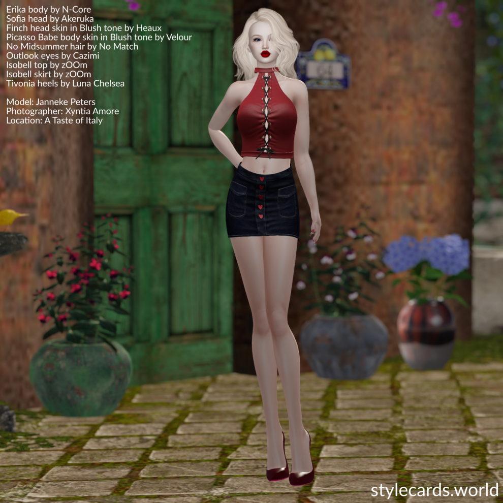 Janneke Peters is standing in front of a home in an Italian village. She has long blonde hair and pale skin and is wearing a red leather halter top, a dark blue denim miniskirt with heart-shaped buttons, and red high heels.